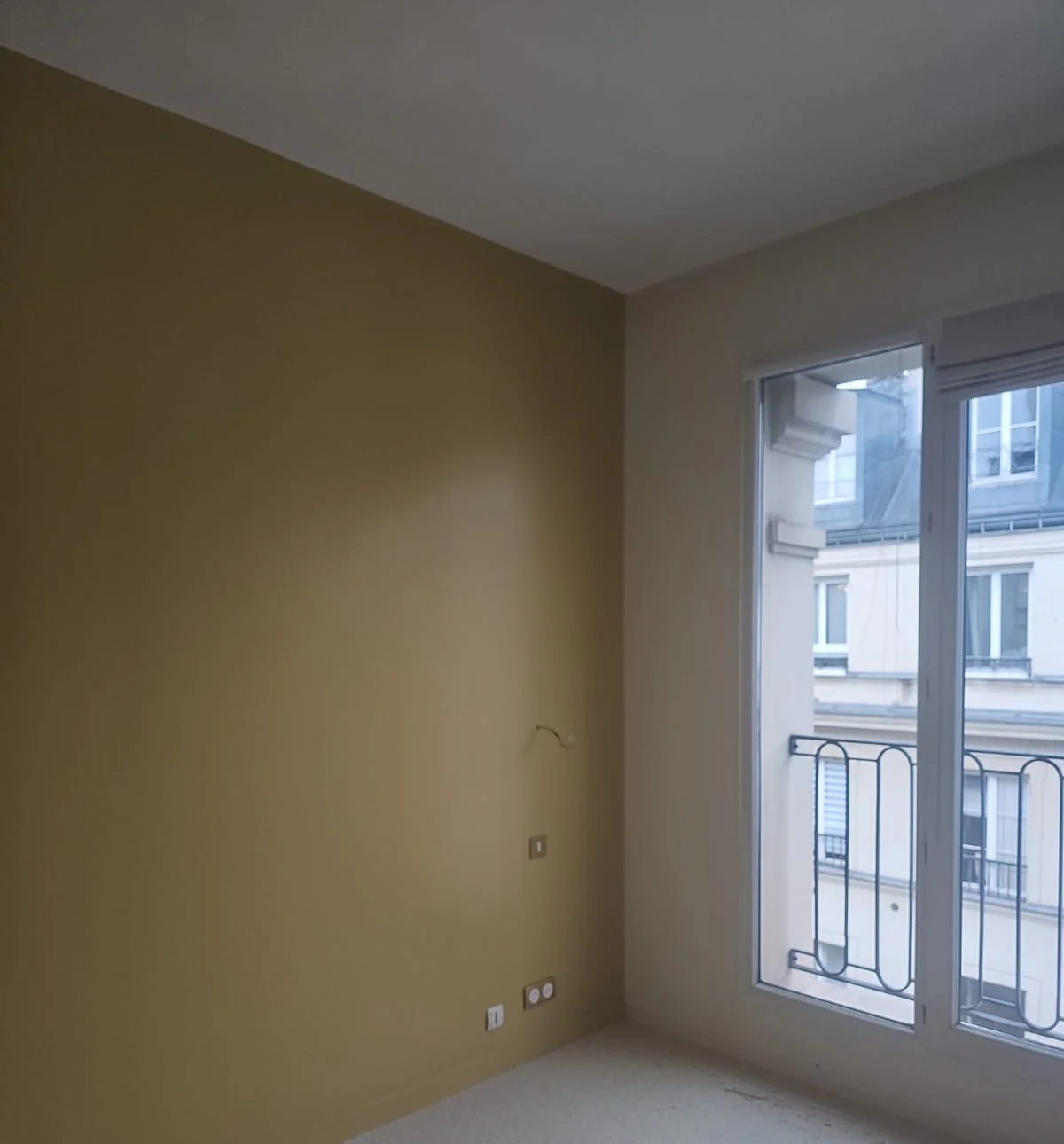 Chambre repeinte avec un mur d'accent jaune moutarde, murs clairs et fenêtre donnant sur une façade haussmannienne