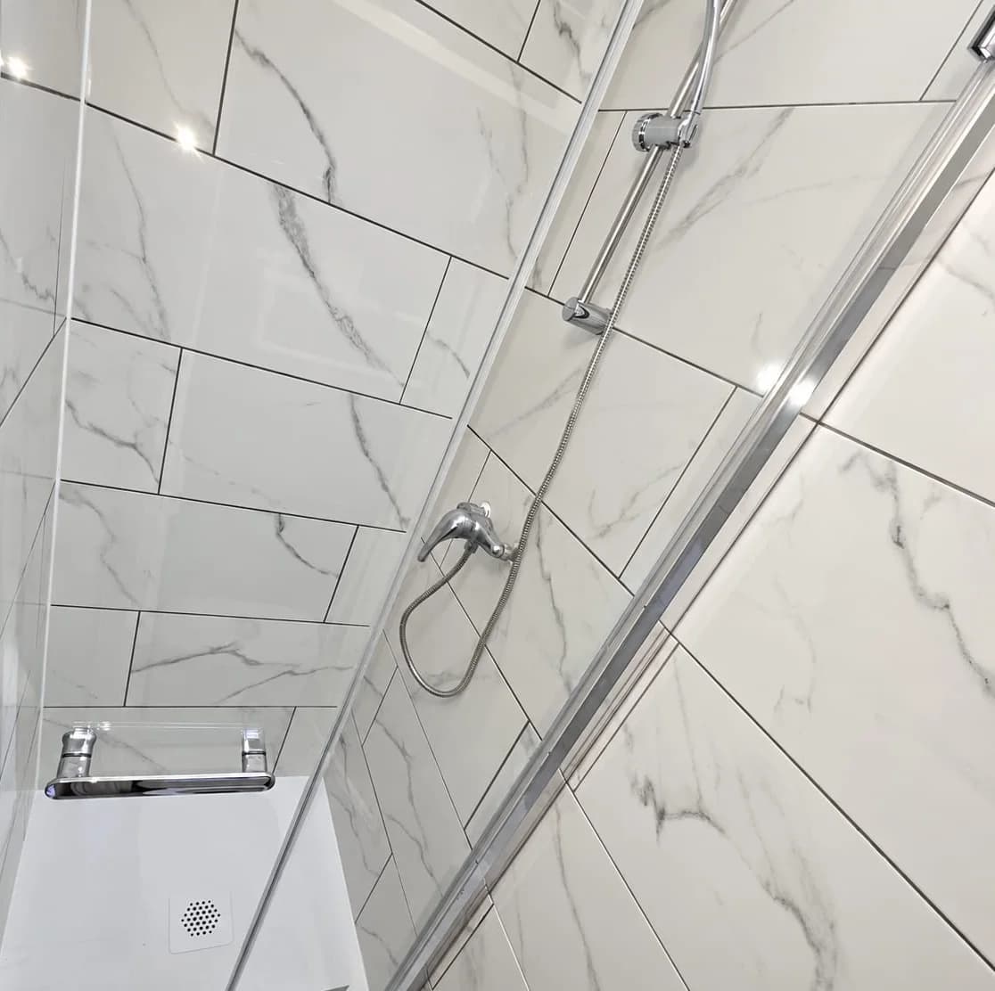 La même douche vue d'ensemble, avec la paroi coulissante en verre, le mitigeur et la douchette sur barre chromée
