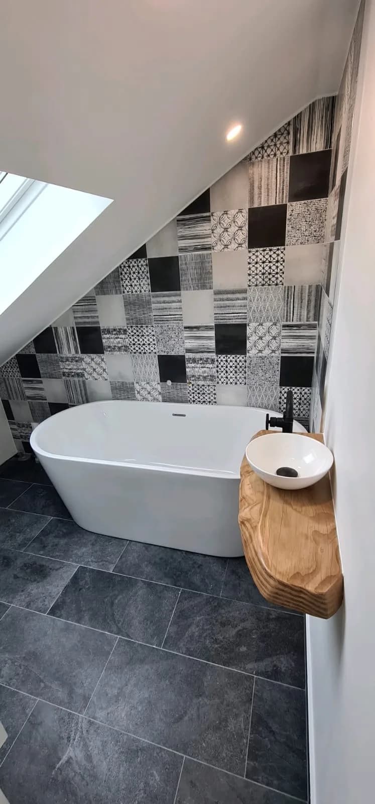 Salle de bain sous combles avec baignoire îlot, faïence à motifs noir et blanc et vasque posée sur une tablette en bois massif